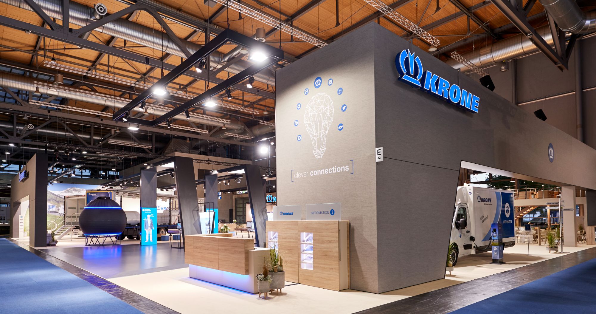 Messestand von Krone auf der IAA in Hannover: grauer Baukörper mit blauem Leuchtschriftzug über dem Eingang, davor eine Holztheke, Vitrinen und ein Transporter unter dem Vordach.