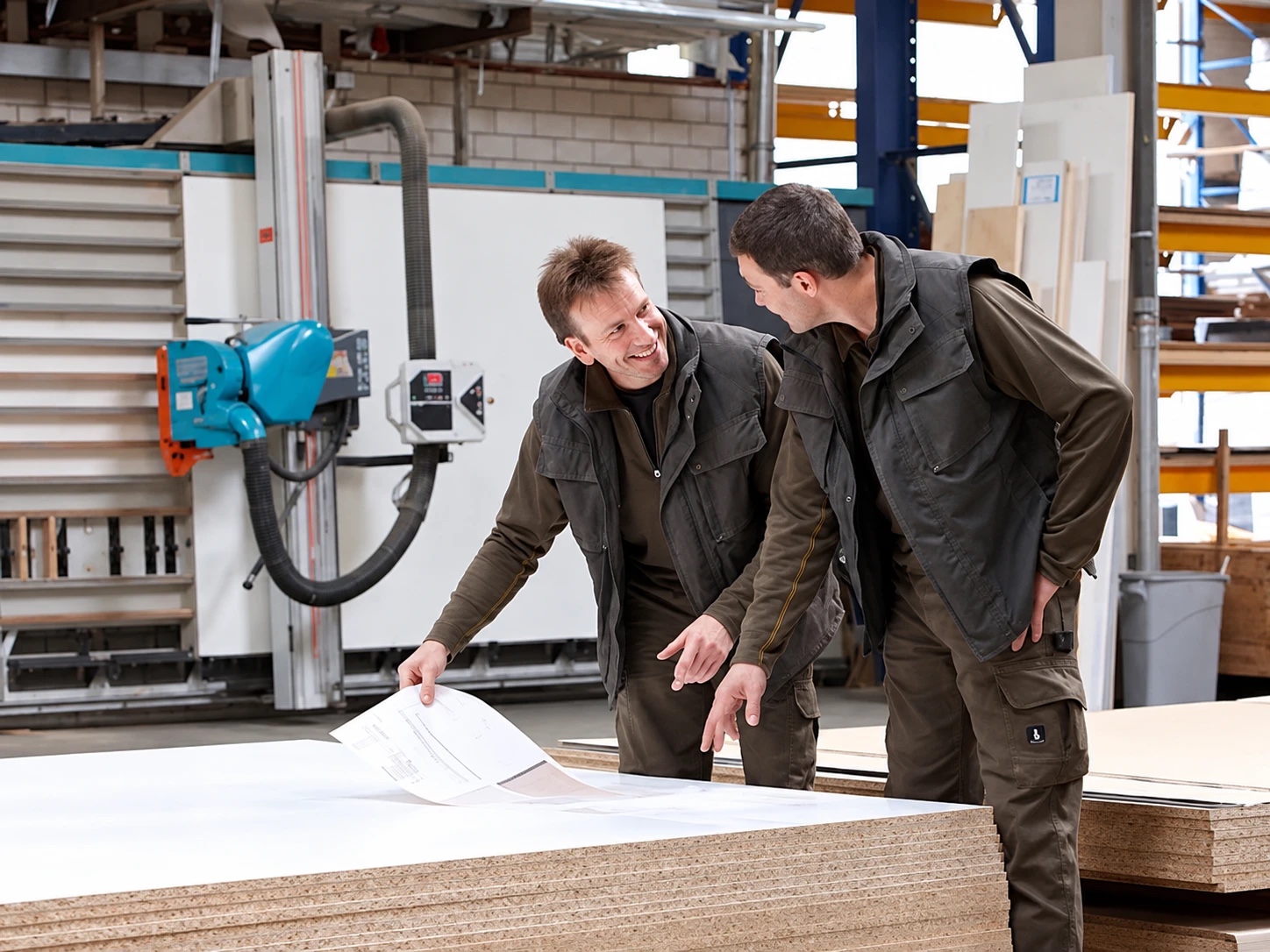 Zwei Mitarbeiter in der eigenen Tischlerei besprechen eine technische Zeichnung über einem Stapel Plattenmaterial, dahinter die stehende Plattensäge.